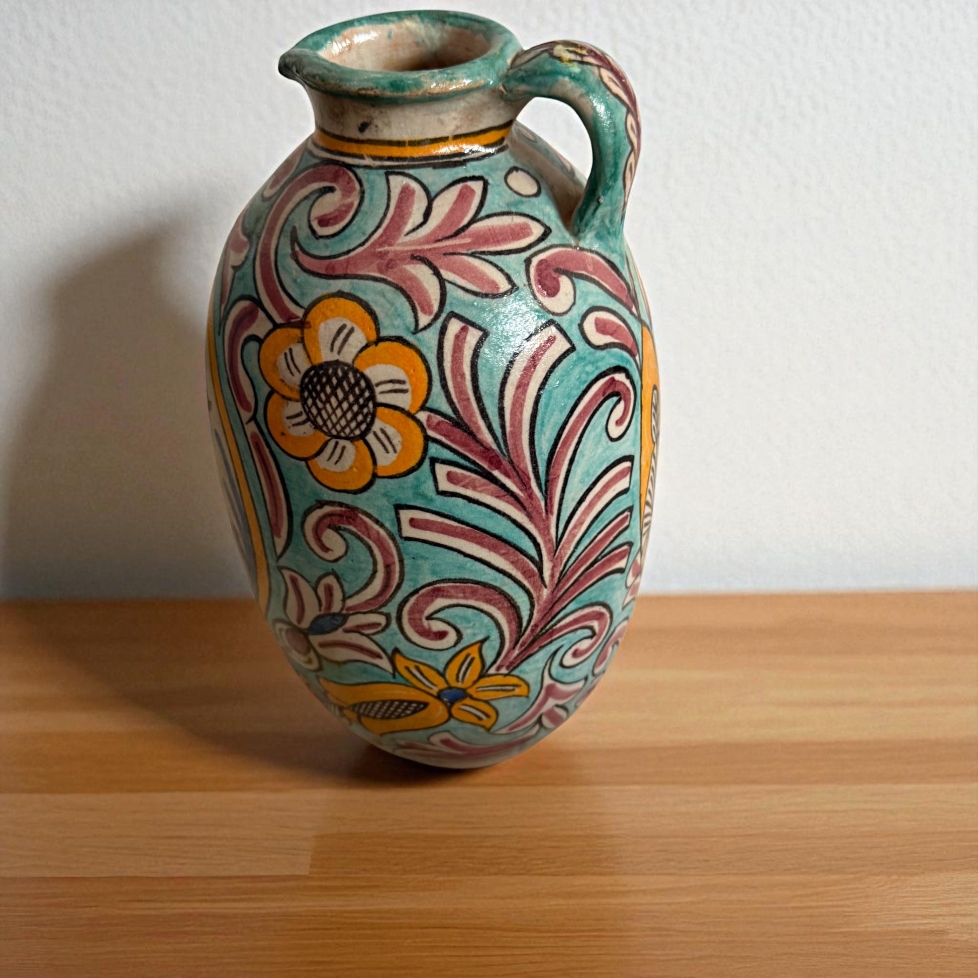 Decorated ceramic pitcher with handle, floral pattern, set on wooden surface.

