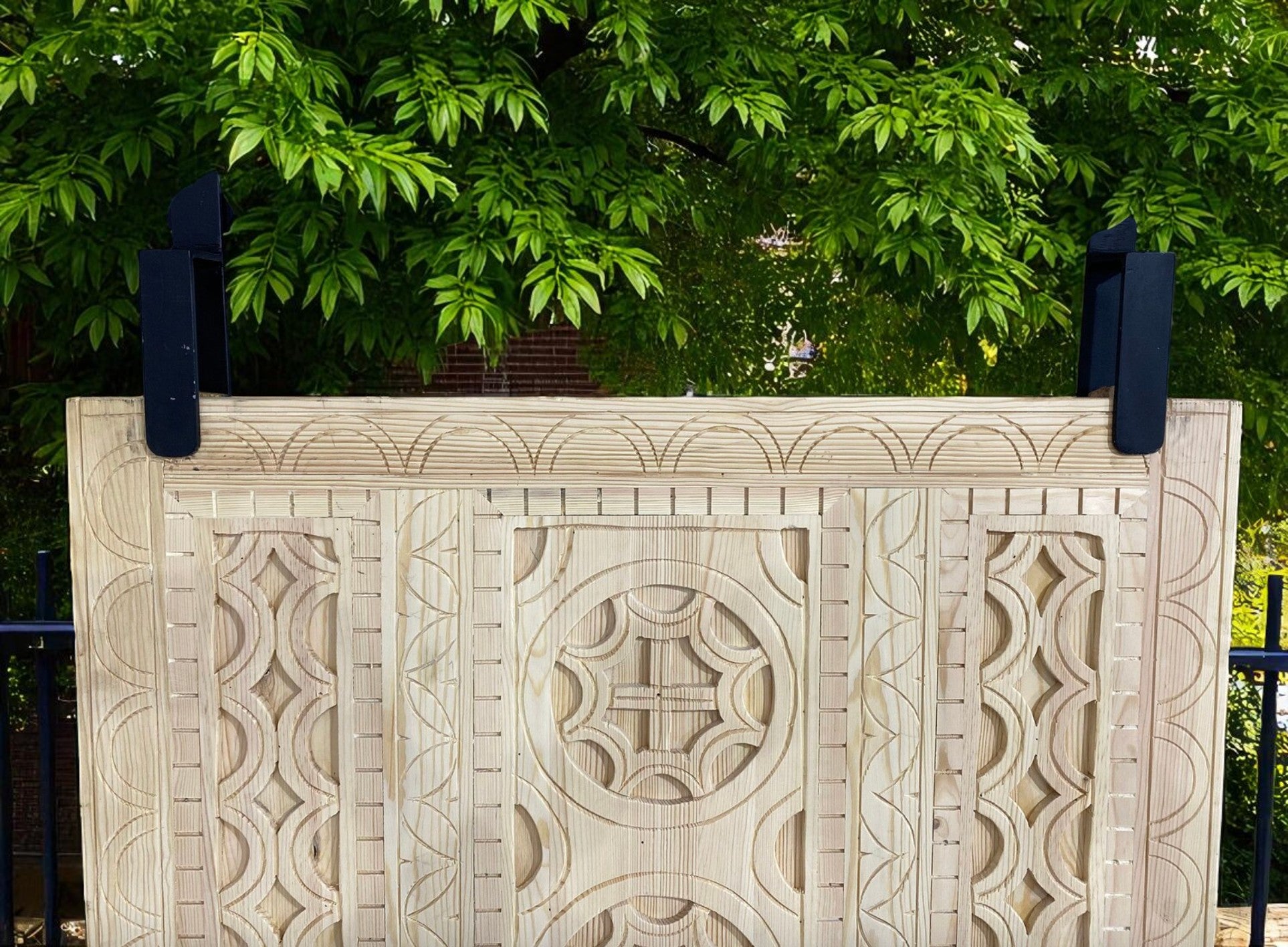 Carved wooden headboard with dark metal supports, green foliage backdrop.