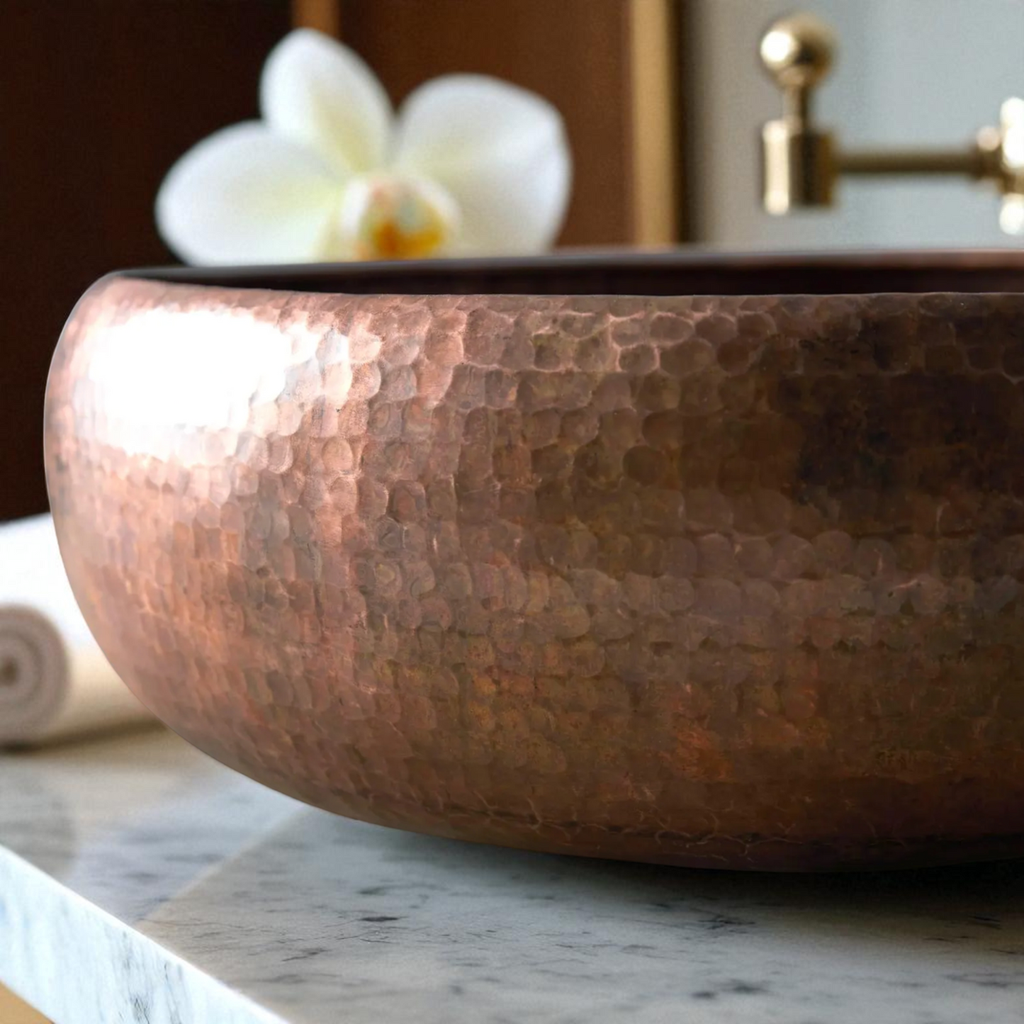Close-up of a hammered copper vessel sink on a marble countertop, with a white orchid and gold faucet in the background.