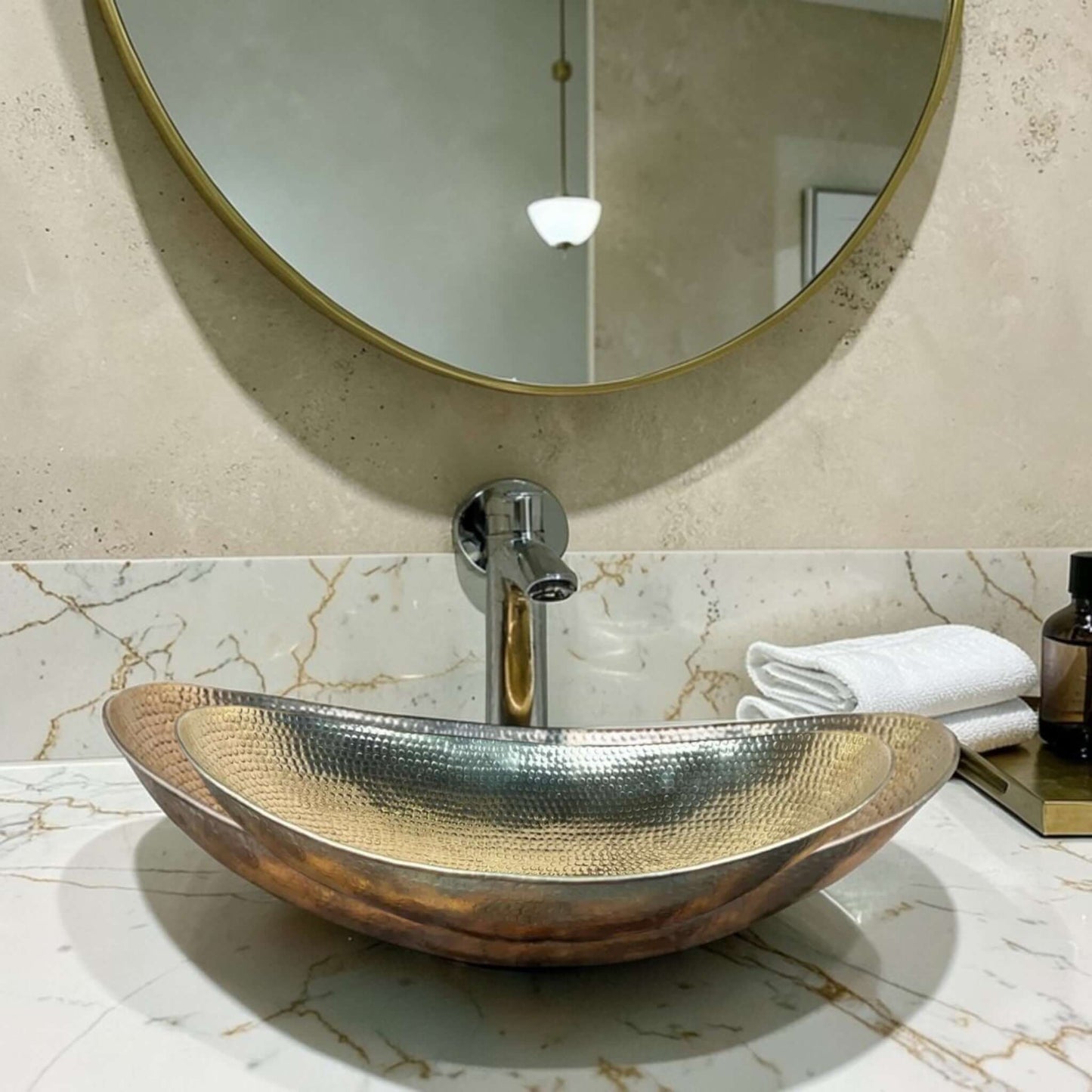 hammered brass bathroom vessel sink displayed on a marble countertop with a round mirror above.