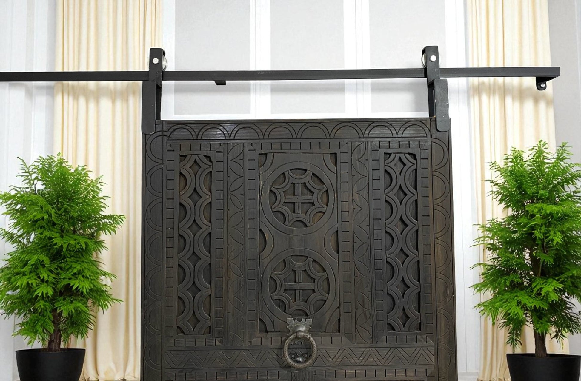 A large, dark wooden door with decorative carvings hangs on a black metal track, flanked by two potted evergreen trees.
