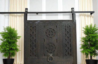 A large, dark wooden door with decorative carvings hangs on a black metal track, flanked by two potted evergreen trees.