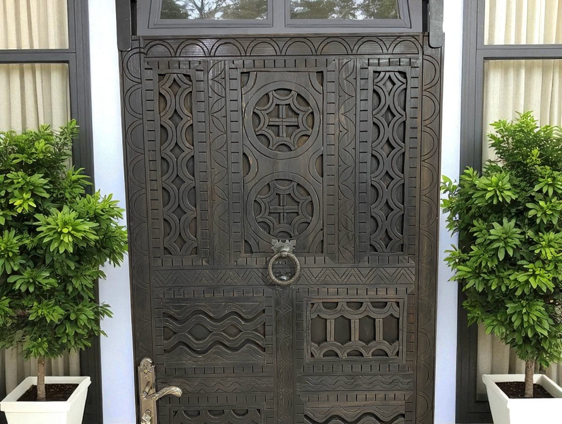 Ornate wooden door with geometric carvings and a ring knocker, flanked by potted trees.