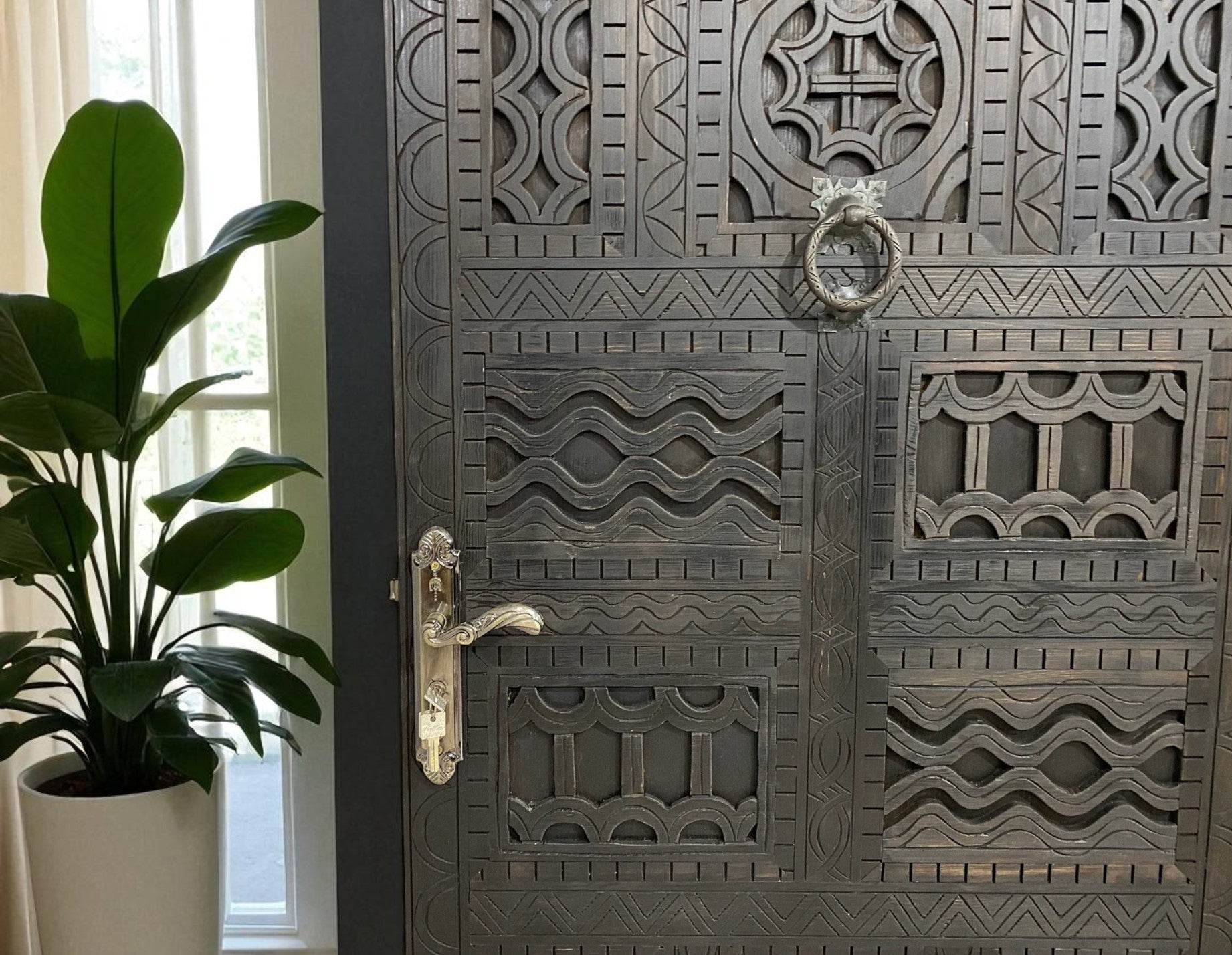 Intricately carved dark wood door with silver handle and knocker, standing next to a large potted plant.

