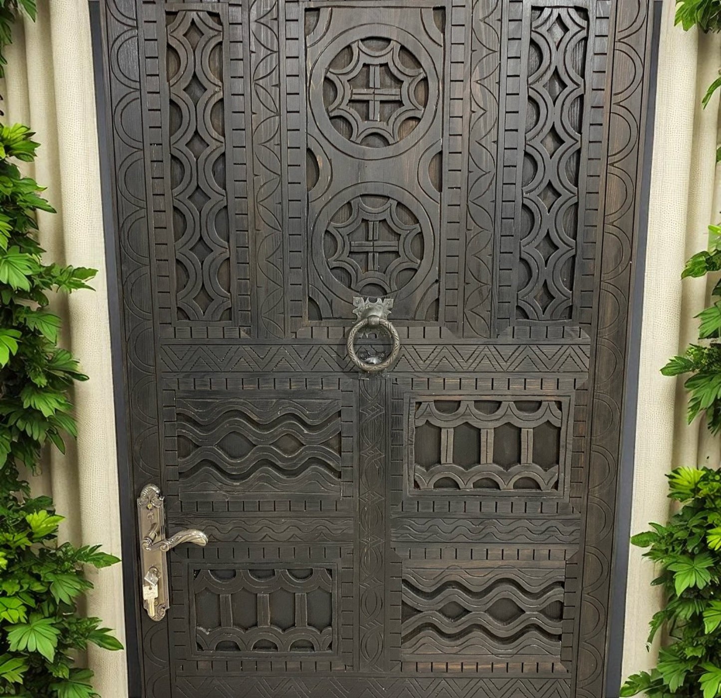 Dark, carved wooden door with geometric patterns, metal door handle, and leafy green plants on both sides.
