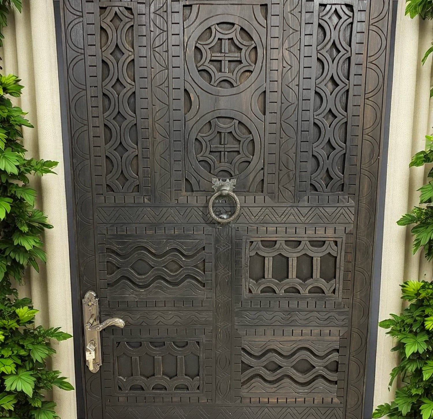 Dark, carved wooden door with geometric patterns, metal door handle, and leafy green plants on both sides.
