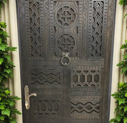 Dark, carved wooden door with geometric patterns, metal door handle, and leafy green plants on both sides.
