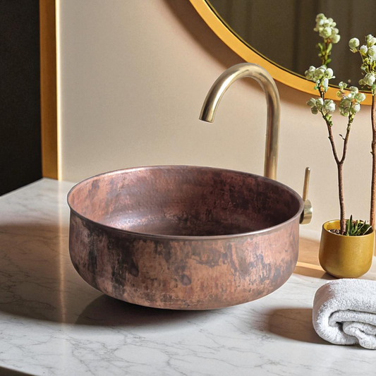 Copper sink with gold faucet and mirror, white towel, and small flower plant.
